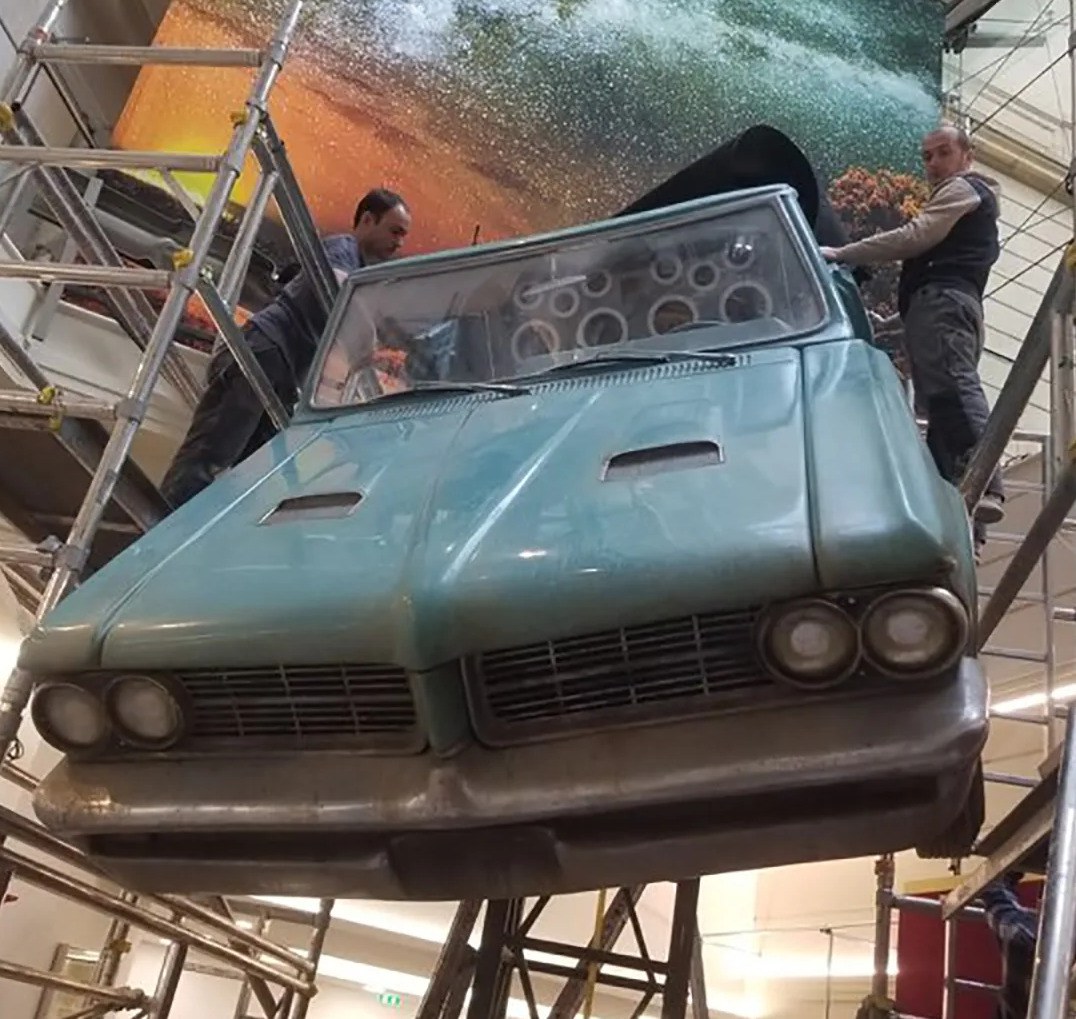 Sub-photo — La voiture est hissée en hauteur devant le décor de fond, sur un mât dessiné pour elle.